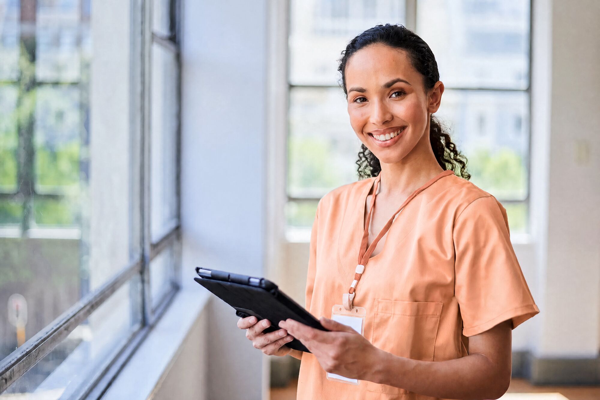 A smiling physician in peach scrubs holding a tablet in a bright clinic